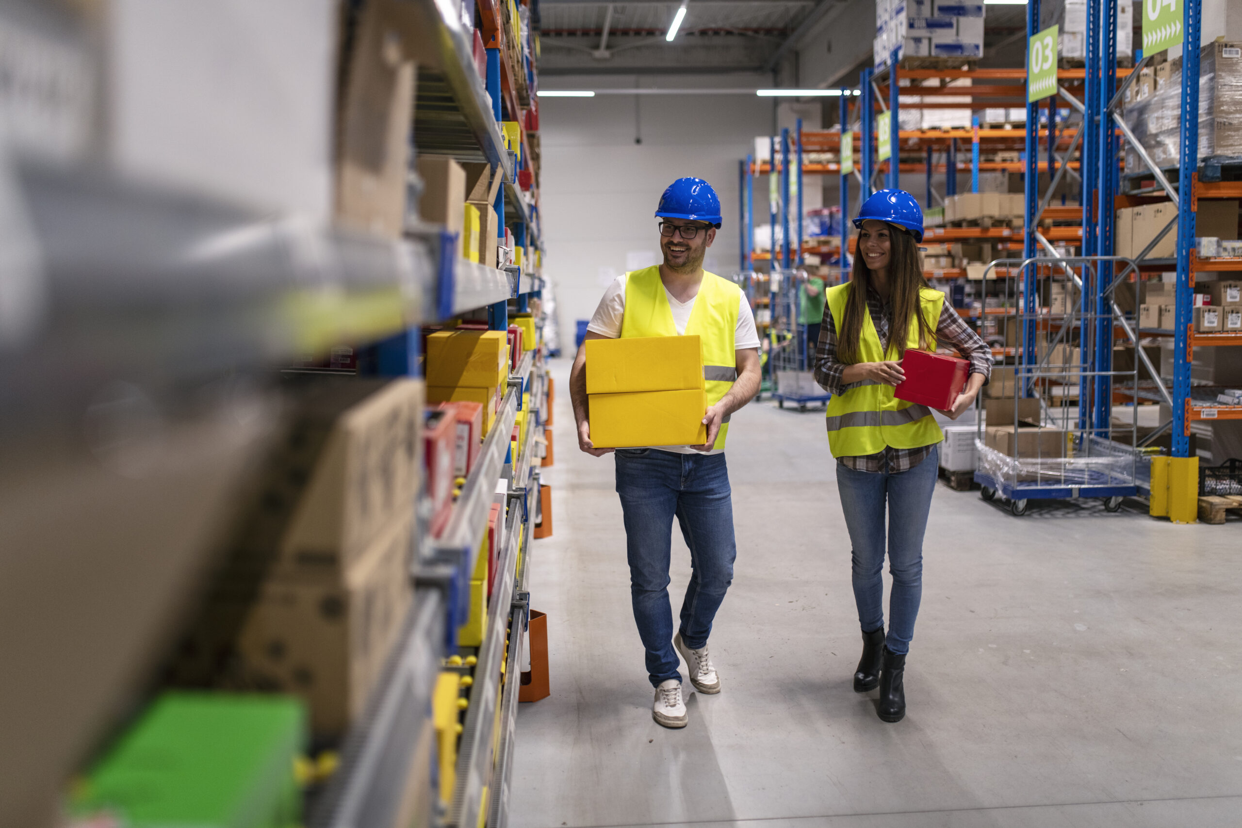 Warehouse workers with helmets and reflective jackets carrying boxes in large distribution center. 16B01UMA10